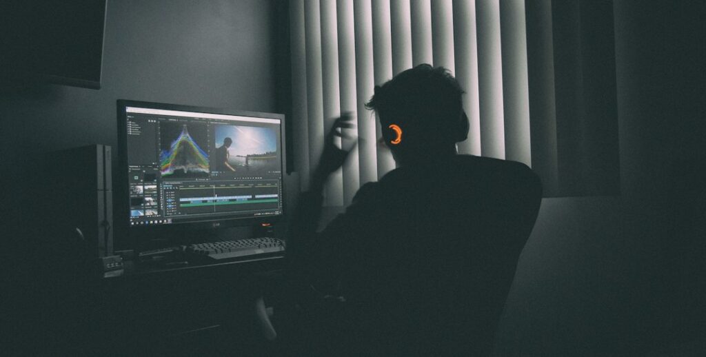 A person in silhouette wearing headphones editing video on a computer in a dark room